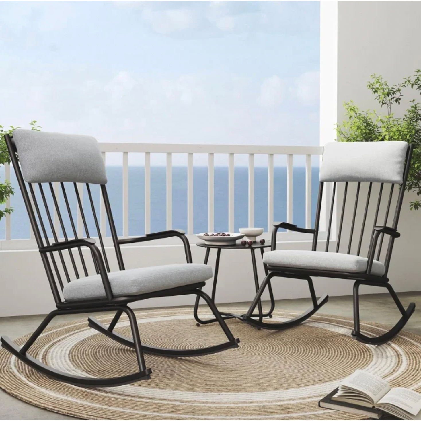 A three-piece patio set featuring two black metal rocking chairs with light gray cushions and a round side table on a woven circular rug, set against a sea view backdrop with a railing and greenery nearby.