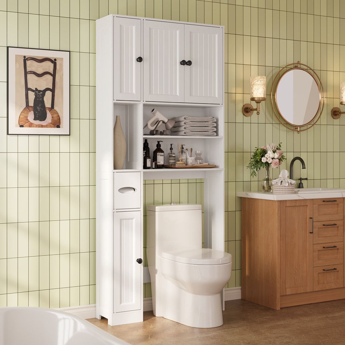Bathroom setup with a white over-the-toilet storage unit featuring shelves and cabinets, a toilet, a wood vanity with a sink, and a round mirror. The green tiled wall includes a chair-themed art piece and wall sconces.