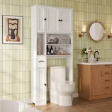 Bathroom setup with a white over-the-toilet storage unit featuring shelves and cabinets, a toilet, a wood vanity with a sink, and a round mirror. The green tiled wall includes a chair-themed art piece and wall sconces.