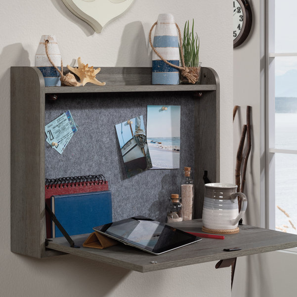 A wall-mounted floating desk with a pinboard, a small shelf, and a fold-down writing surface, styled with a nautical theme including decorative bottles, buoys, and a starfish.