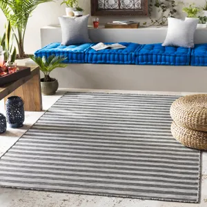 A 6' x 9' Breakwater Bay Striped Rug featuring gray and white horizontal stripes is displayed in a cozy room with a blue cushioned bench, woven poufs, decorative plants, and lanterns.
