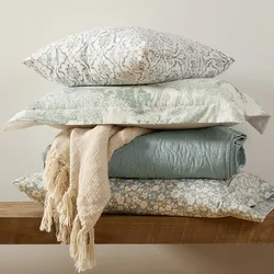 A neatly stacked arrangement of printed and textured pillows, a woven beige blanket, and a folded quilt in soft, pastel colors sits on a wooden surface.