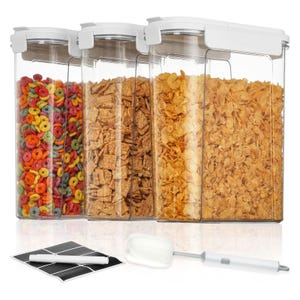 Clear airtight cereal storage containers holding colorful loops, cinnamon squares, and corn flakes, with a cleaning brush and chalkboard labels with a marker.