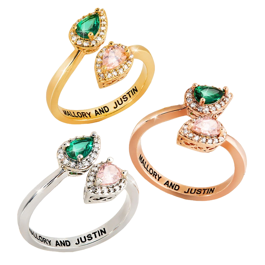 The Couple's Custom Birthstone Ring features a split shank design with two pear-shaped gemstones, one green and one pink, each surrounded by small clear stones. The band can be personalized with names, as shown with \“Mallory and Justin,\“ and is available in gold, silver, and rose gold finishes.