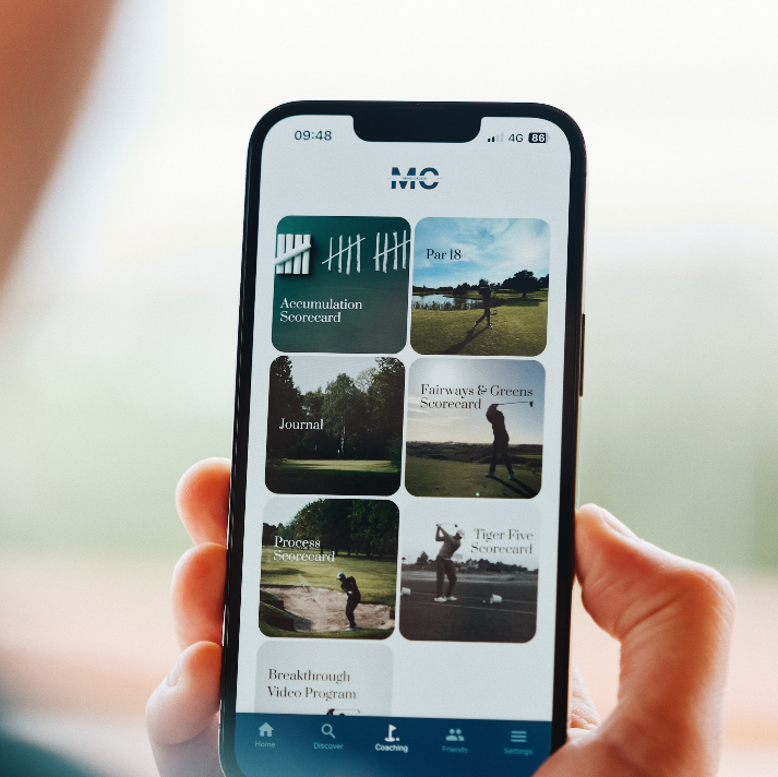 A person holds a smartphone displaying a golf coaching app with options like Accumulation Scorecard, Par 18, Journal, Fairways & Greens Scorecard, Process Scorecard, Tiger Five Scorecard, and Breakthrough Video Program.