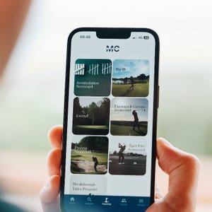 A person holds a smartphone displaying a golf coaching app with options like Accumulation Scorecard, Par 18, Journal, Fairways & Greens Scorecard, Process Scorecard, Tiger Five Scorecard, and Breakthrough Video Program.