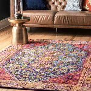 A colorful, intricately patterned Bohemian rug with vibrant reds, blues, and yellows lies on a wooden floor, paired with a brown leather sofa and a small round gold side table.