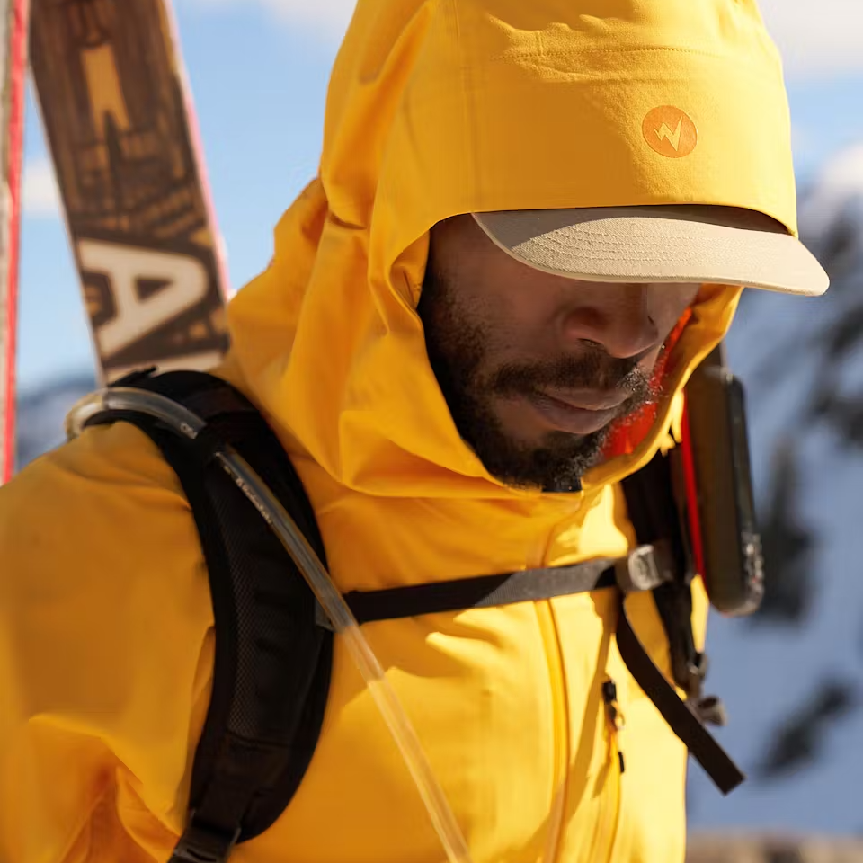 Yellow Marmot Refuge Pro Ski Jacket with a hood, featuring a hydration tube on the chest and a backpack strap, designed for skiing.