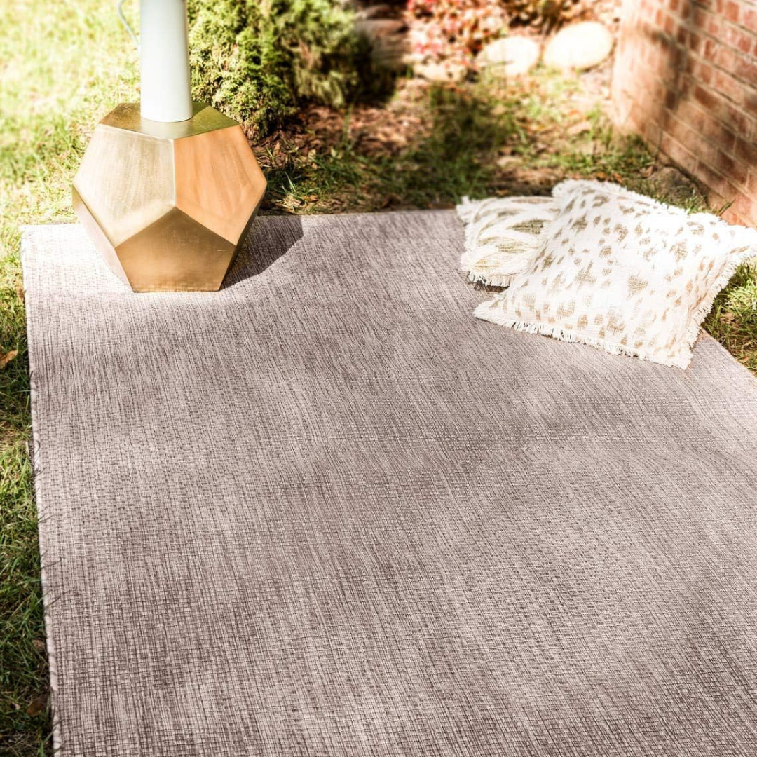 A gray outdoor rug is spread on the grass with a geometric gold side table and two decorative fringed pillows.