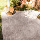 A gray outdoor rug is spread on the grass with a geometric gold side table and two decorative fringed pillows.