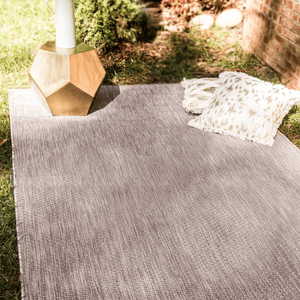A gray outdoor rug is spread on the grass with a geometric gold side table and two decorative fringed pillows.