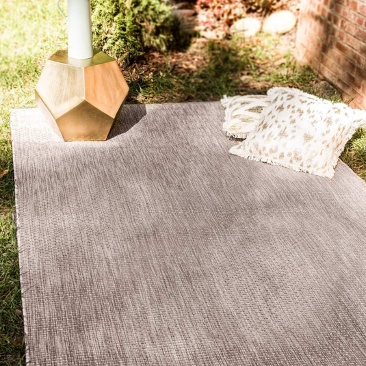 A gray outdoor rug is spread on the grass with a geometric gold side table and two decorative fringed pillows.