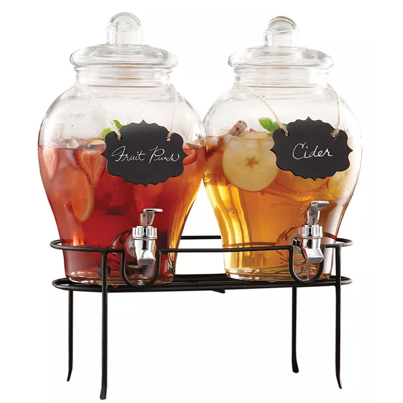 Set of two glass beverage dispensers with clear lids, labeled \“Fruit Punch\“ and \“Cider,\“ each featuring a metal spigot, held on a black metal stand.