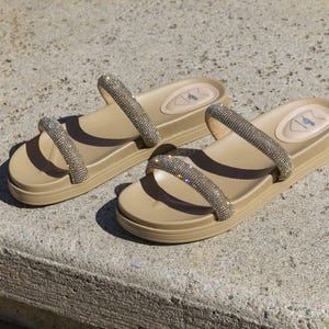Beige flat sandals with two rhinestone-encrusted straps and a slightly raised sole.