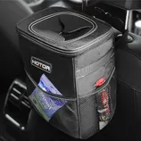 A black car trash can with a flip-top lid is hanging on the back of a car seat, featuring side mesh pockets and a white and red logo on the front.