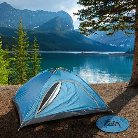 A blue camping tent set up by a lake with mountains in the background; a blue frisbee lies in the foreground.