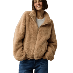 A woman is wearing a tan zip-up sherpa jacket with a high collar and elastic cuffs, paired with blue jeans. The jacket has a cozy, fluffy texture and a relaxed fit.