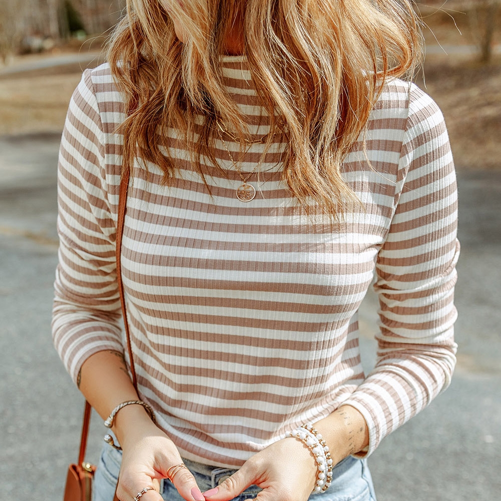 The top is a long-sleeve, fitted shirt with horizontal beige and white stripes and a crew neckline, accessorized with a small round pendant necklace.