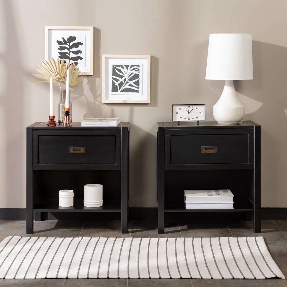 Two black bedside tables with a drawer and shelf, featuring decorative items like candles, white containers, a book, and a white lamp. Art prints in light frames and a small clock adorn the wall above. A striped rug lies in front.