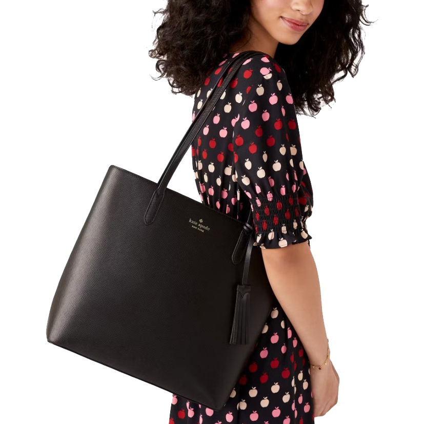 A woman carries a black Kate Spade tote bag with a tassel detail, wearing a dress patterned with red, pink, and white apples.