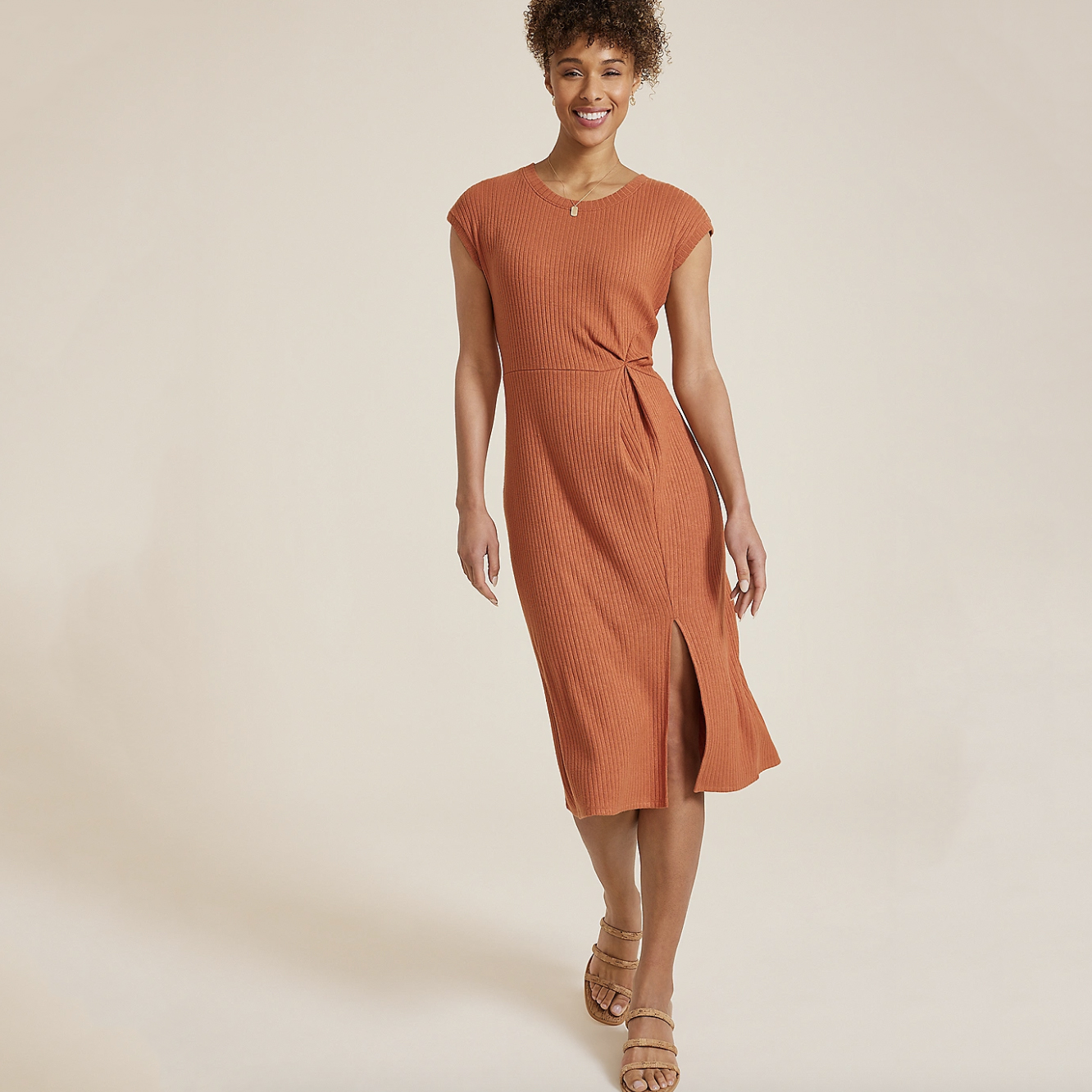 A woman is wearing an orange, ribbed, knee-length dress with a twist detail at the waist, paired with brown strappy sandals.