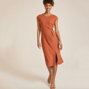 A woman is wearing an orange, ribbed, knee-length dress with a twist detail at the waist, paired with brown strappy sandals.