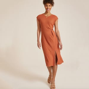 A woman is wearing an orange, ribbed, knee-length dress with a twist detail at the waist, paired with brown strappy sandals.