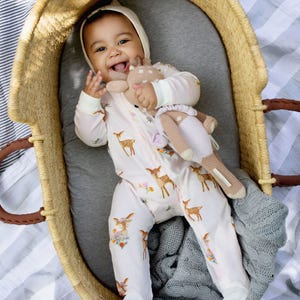 A baby wearing a deer-patterned onesie lies in a wicker basket, holding a knitted toy. A gray knitted blanket provides additional comfort.