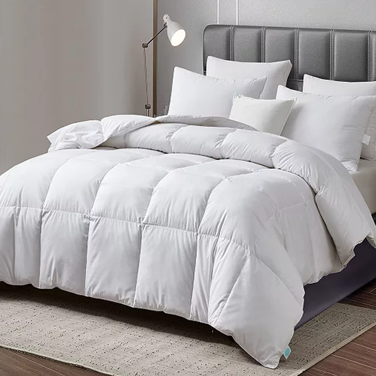 A white quilted goose feather and down comforter is neatly spread on a queen-sized bed, accompanied by a set of matching pillows. The comforter has a soft and plush appearance, providing a cozy and inviting look.