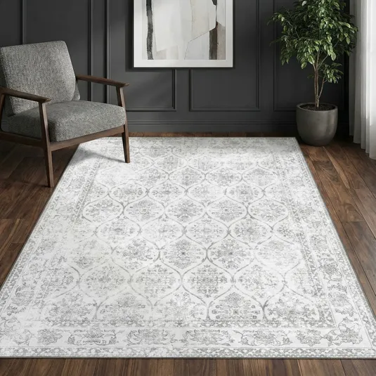 A patterned area rug with a floral design is placed on a wooden floor, accompanied by a gray upholstered chair, a potted plant, and abstract wall art in the background.