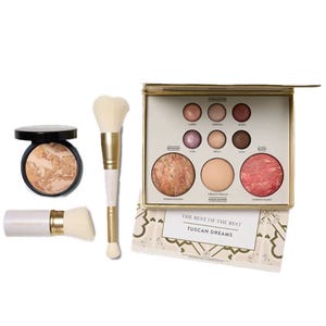 A makeup collection including a palette with various baked eyeshadows and blushes, a powder compact, and two fluffy makeup brushes, all themed \“Tuscan Dreams.\“