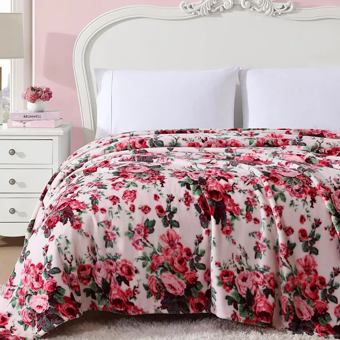 A plush floral blanket with a pattern of red roses and green leaves on a light pink background, displayed on a white bed with a decorative headboard, accompanied by white pillows and a bedside table with books and flowers.