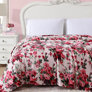 A plush floral blanket with a pattern of red roses and green leaves on a light pink background, displayed on a white bed with a decorative headboard, accompanied by white pillows and a bedside table with books and flowers.