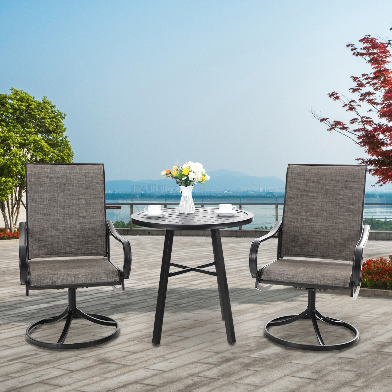 A 3-piece patio bistro set featuring a round table with a slatted top and a vase of flowers, accompanied by two swivel chairs with gray textilene mesh seats and black metal frames, set on a paved outdoor area with a scenic view.