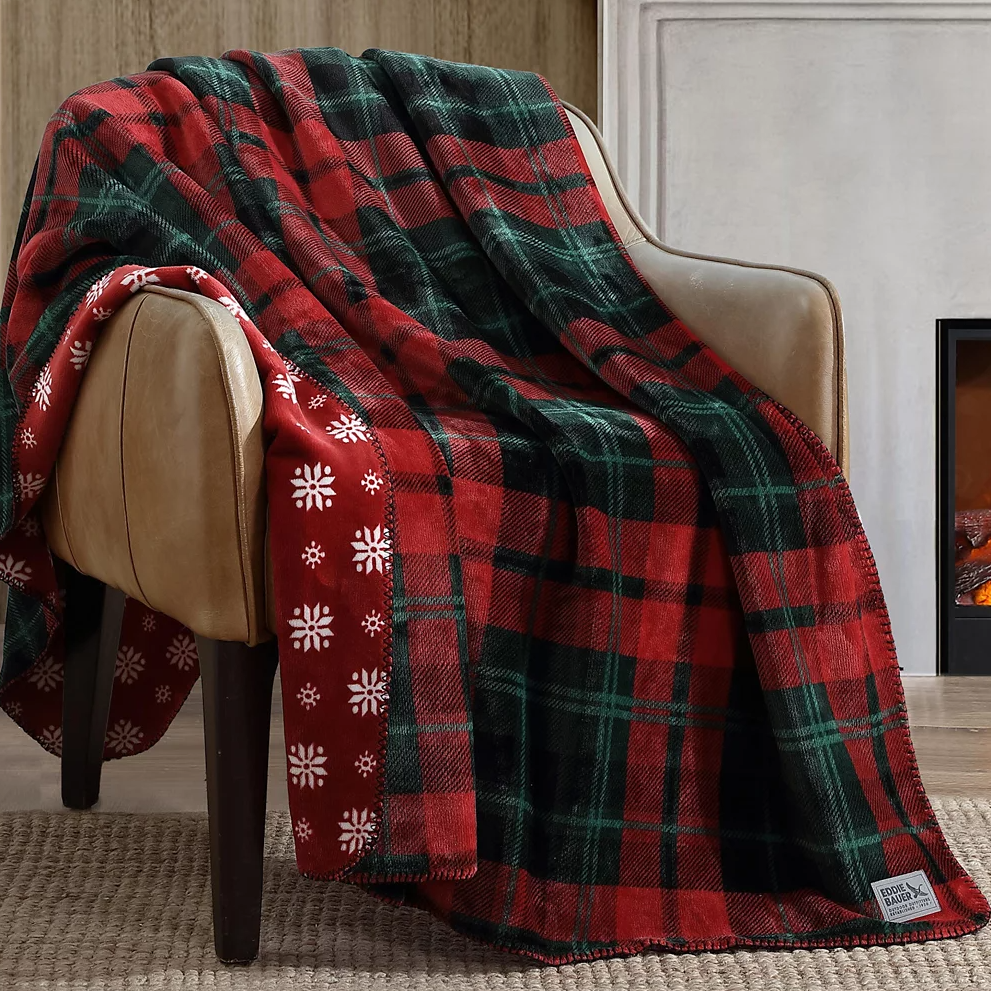 A cozy plaid blanket with red and green colors draped over a leather armchair. It features a snowflake design on the reverse side.