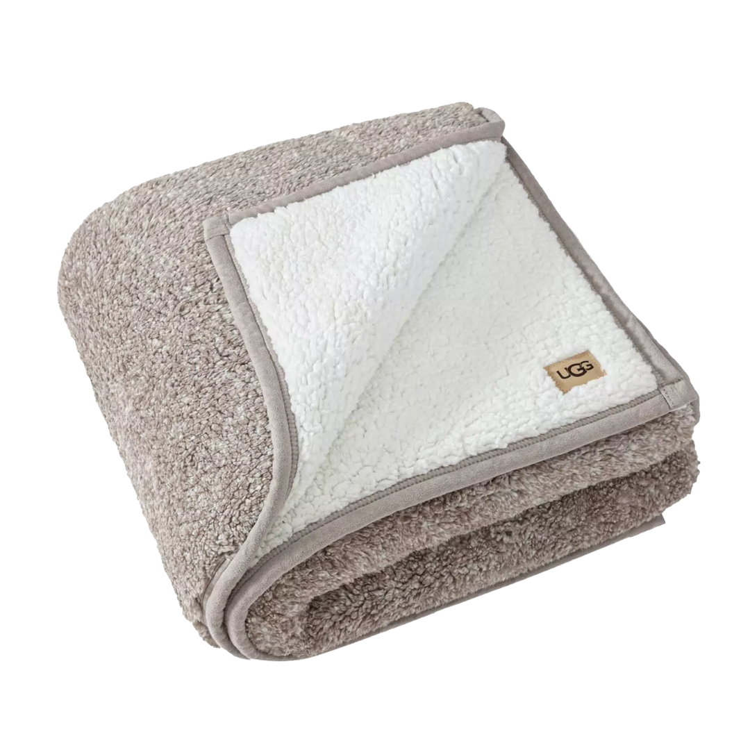 A cozy, folded blanket with a textured gray outer layer and a soft white sherpa lining, featuring a small \“UGG\“ logo tag on the corner.