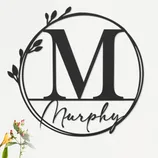 A decorative wall art piece featuring the letter \“M\“ in a circle with botanical accents, accompanied by the name \“Murphy,\“ displayed above a sofa with flowers nearby.
