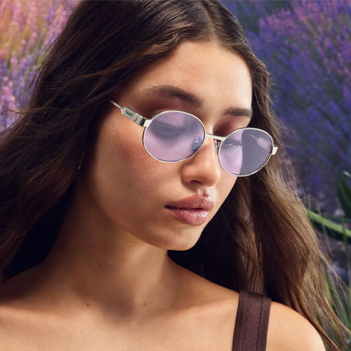 A woman wearing silver-framed sunglasses with round, pink-tinted lenses is shown against a backdrop of purple flowers.