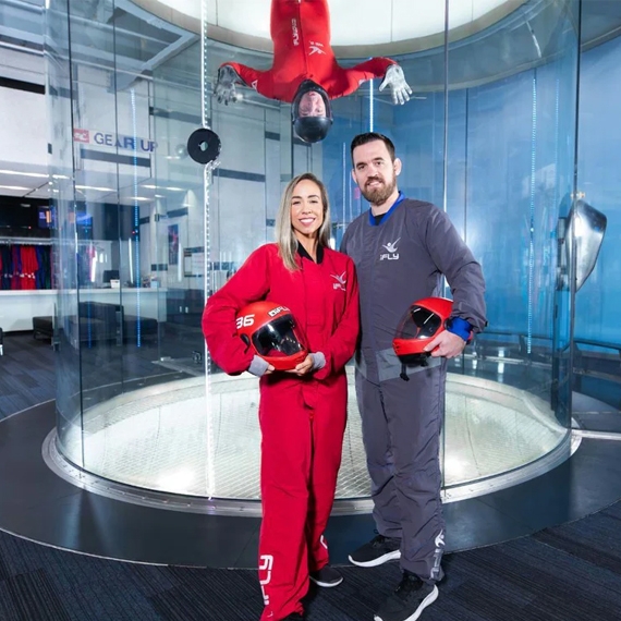 Two people in indoor skydiving suits hold helmets, standing in front of a wind tunnel, while another person is flying upside down inside the tunnel.