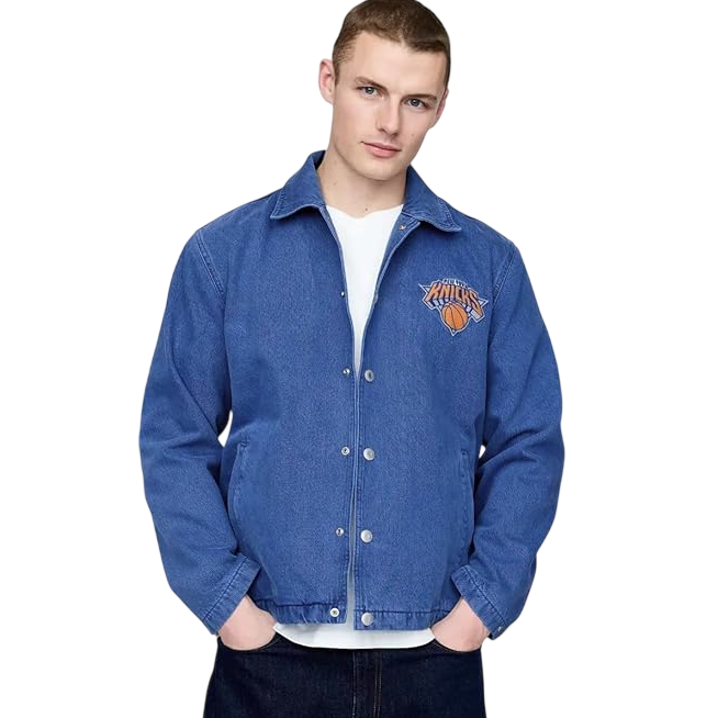 A person wears a blue button-up jacket featuring the New York Knicks logo, over a white shirt and dark jeans.