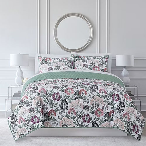 A bed with a floral-patterned comforter and matching pillow shams, topped with a solid green blanket, is flanked by two white lamps on matching side tables. A round mirror hangs above the bed.