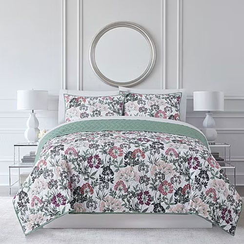 A bed with a floral-patterned comforter and matching pillow shams, topped with a solid green blanket, is flanked by two white lamps on matching side tables. A round mirror hangs above the bed.