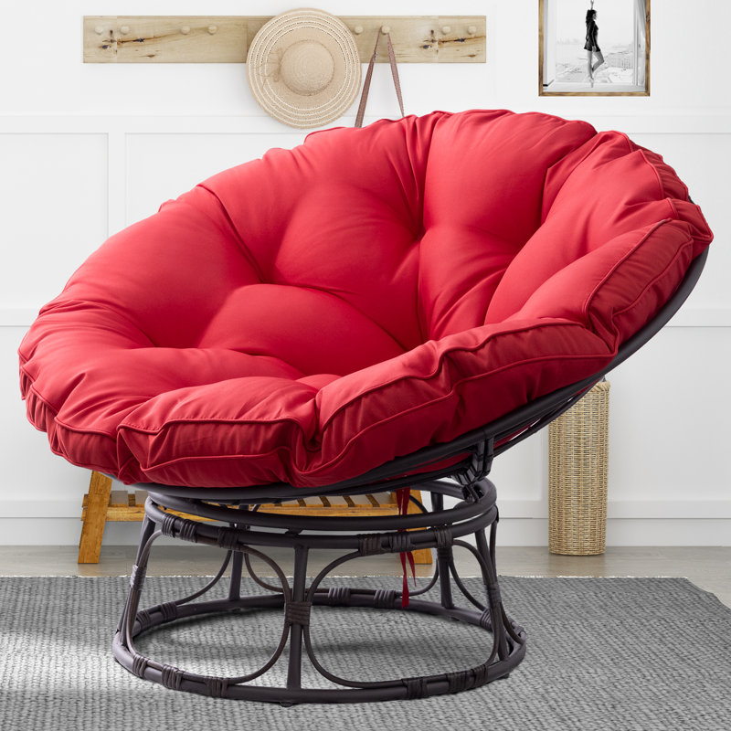 A red-cushioned swivel Papasan chair with a round, tufted seat, supported by a black rattan frame. The chair is set on a gray rug in a bright room with a straw hat and framed photo in the background.