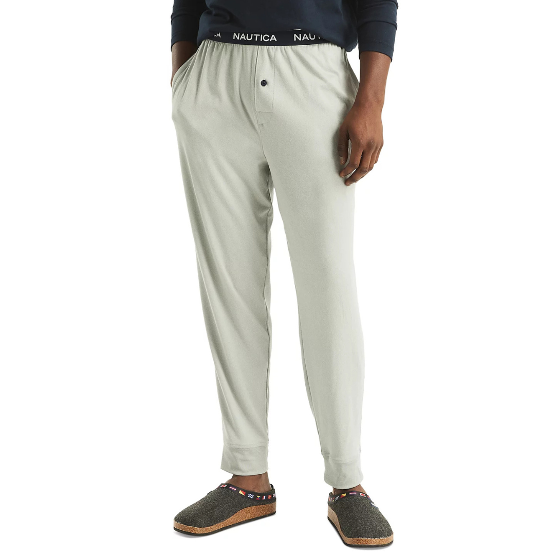 Men's pale gray pajama pants with an elastic waistband featuring the \“Nautica\“ logo and a button fly, paired with gray wool slippers with multicolored strap detailing.