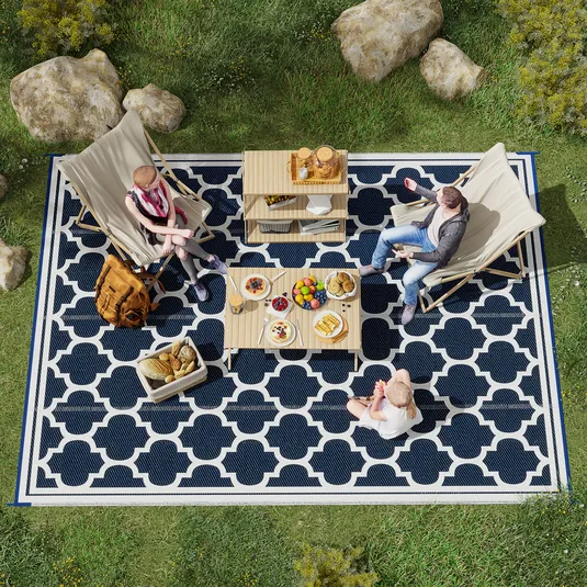 Outdoor scene with a blue and white patterned rug, two wooden tables, foldable chairs, and a variety of food including fruits and pastries.