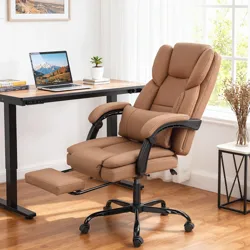A brown ergonomic office chair with a retractable footrest is placed next to a wooden desk with a laptop and a small plant in a bright, modern room.