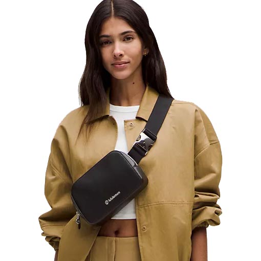 A person wears a tan shirt and pants with a small black crossbody bag featuring a wide strap and buckle.