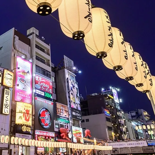 The scene shows a vibrant street with illuminated lanterns and colorful billboards, including advertisements for ramen restaurants and other eateries, set against a night sky backdrop.