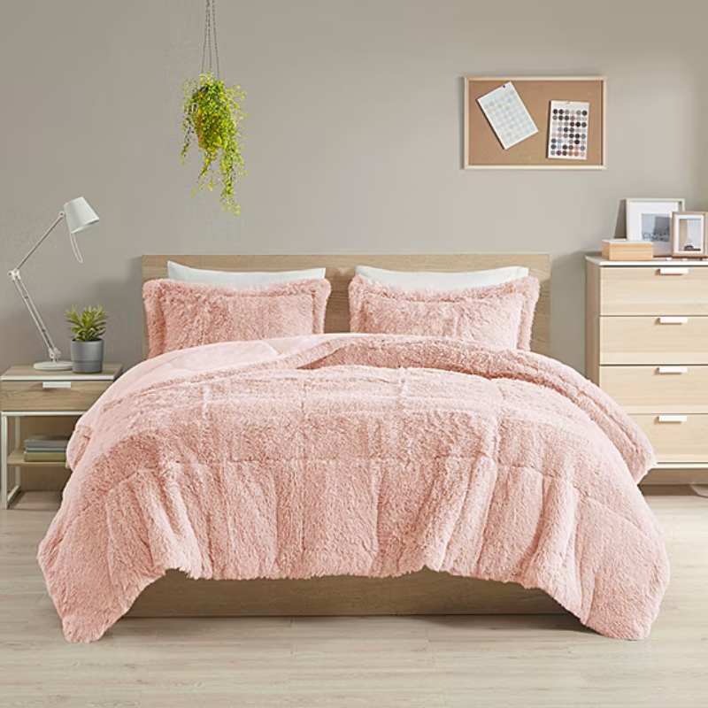 A cozy bedroom features a bed with a fluffy pink comforter and matching pillows. The room includes a wooden headboard, nightstand with a lamp, a hanging plant, and dresser with framed photos, creating a warm and inviting atmosphere.
