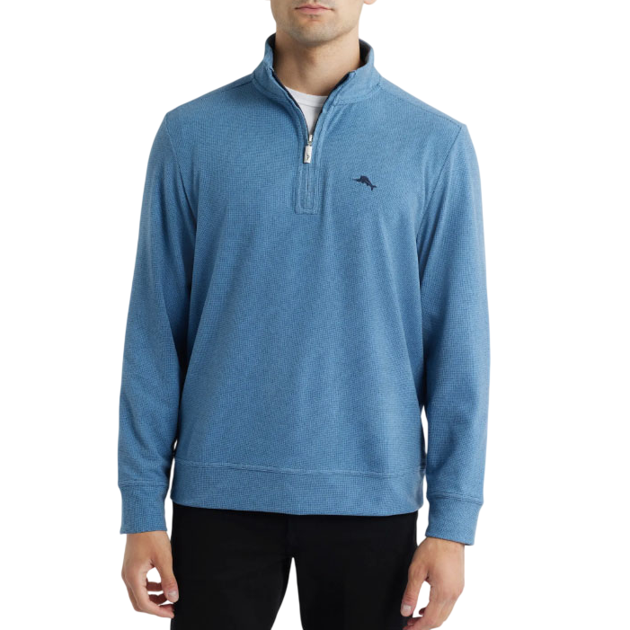 A man is wearing a blue quarter-zip pullover sweater with a small embroidered logo on the chest, paired with black pants.